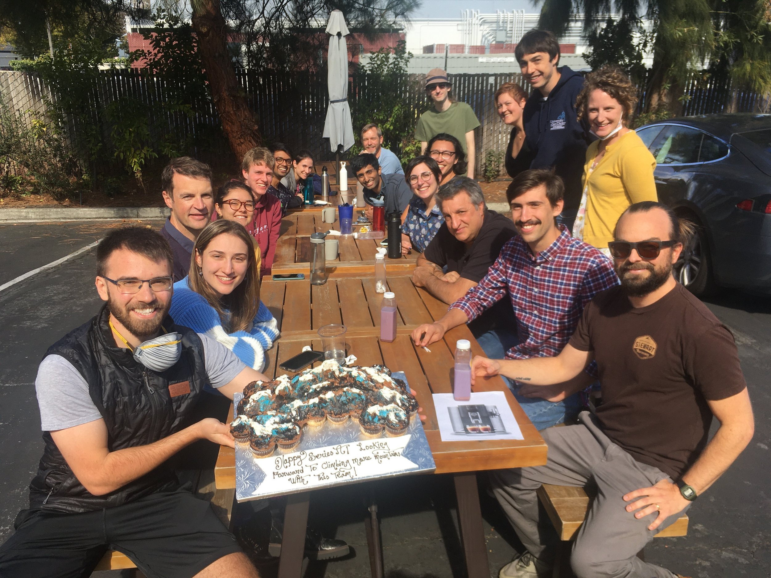 Group of several individuals sitting around a picnic table outside. There's a cake on the table that reads "Happy Series A. Looking forward to climbing more mountains with this team."